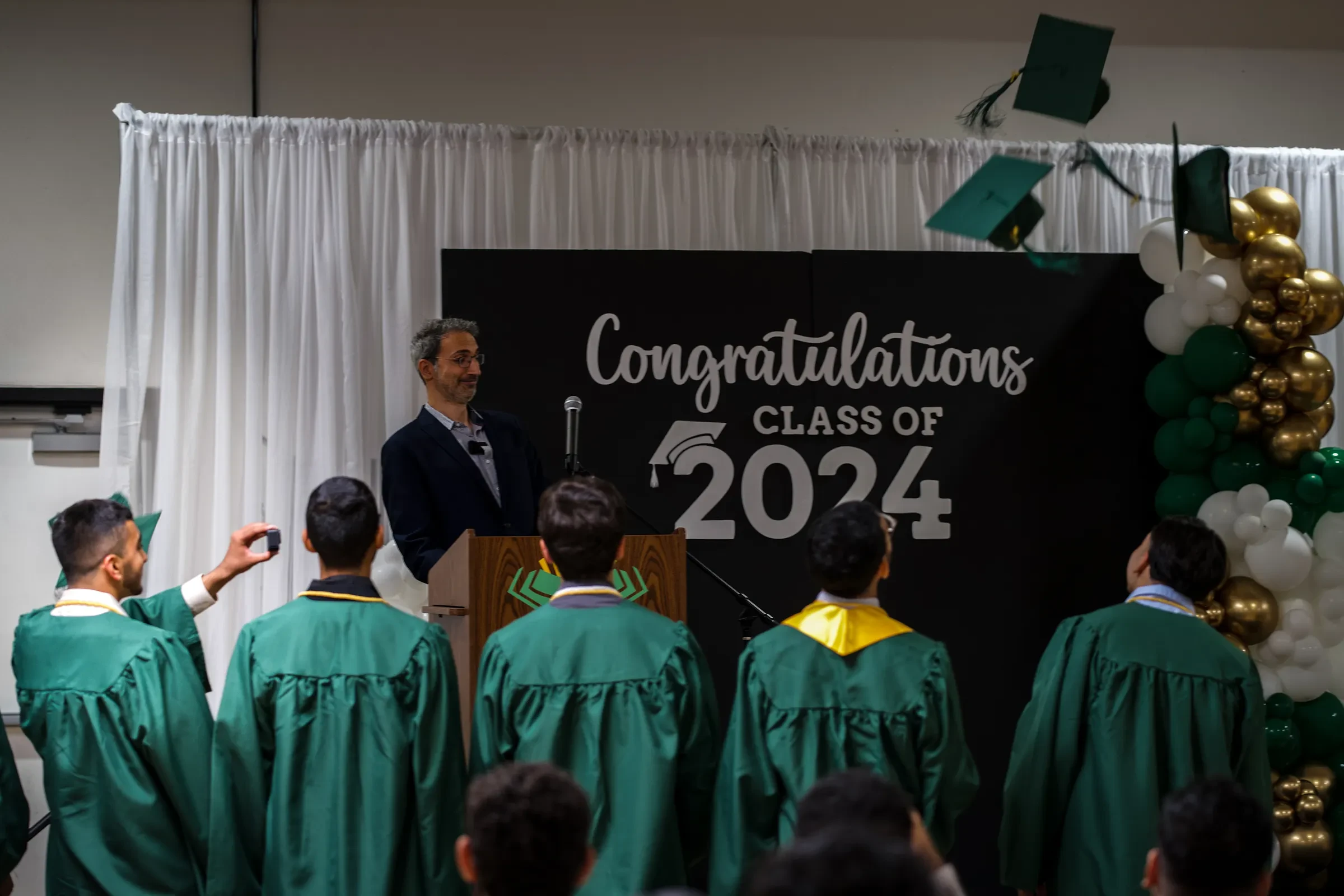 Graduates in green gowns facing a “Congratulations, Class of 2024” backdrop.